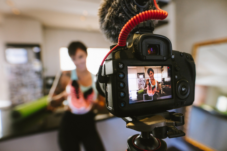 Repurpose Content Concept - Female vlogger holding sports shoes in hands happily looking in camera while recording video for blog. Focus on camera with social media influencer reviewing and marketing a sports shoe